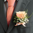 A single peach rose, perfectly formed and softly blushed, is the centrepiece of this handcrafted boutonniere, resting with confident ease on a charcoal suit lapel. Its petals are plush and warm, a coral-pink that deepens toward the centre, reminiscent of dawn light falling across the Gerrards Cross High Street on an early summer morning. Tiny sprays of white baby's breath (gypsophila) cluster around the rose like a lace halo, their fine texture offering a whimsical counterpoint to the rose's velvet surface. Wisps of fresh green foliage and a slender curled leaf provide structure and a hint of woodland energy-calling to mind nearby Burnham Beeches and the dew-sparkle of local hedgerows. The base is bound with a satin ribbon in matching coral-pink, tied into a tidy bow that mirrors the tone of the soft pink tie beneath the lapel; the ribbon's sheen adds a polished finish without overwhelming the bloom. Against the suit's charcoal fabric, colours are sharpened and given depth, each element rendered with clarity and calm. There is a faint, gentle scent to the arrangement-sweet, slightly citrus-tinged rose notes with the green freshness of cut foliage-that suggests celebration and hopeful beginnings. Prepared with the attention of an experienced florist for weddings, anniversaries, or milestone celebrations, this boutonniere is something Flower Delivery Gerrards Cross would carefully pin for a groom or cherished guest, reflecting the town's refined, welcoming character and the quiet craft of local floristry.