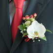 This is a thoughtfully arranged boutonnière photographed close-up on the left lapel of a tailored dark charcoal suit, a composition that reads both modern and timeless in equal measure. The centerpiece is a pristine white Phalaenopsis orchid whose broad petals spread with calm confidence; under soft daylight the bloom appears almost luminous, and the orchid's yellow-gold patterned lip adds a precise, painterly detail at its core. Surrounding foliage of lustrous green leaves provides a grounding backdrop, their sheen contrasting with the orchid's satiny surface and the matte wool of the jacket. Delicate sprays of gypsophila float like tiny stars across the arrangement, imparting a sense of air and celebration, while a cluster of warm pink-red hypericum berries introduces a rounded texture and a complementary blush that repeats the tone of the nearby solid red satin tie. A narrow loop of matching red ribbon ties the stems at the base, a florist's finishing touch that mirrors the tie's sheen and creates visual harmony. The cuff of a light patterned dress shirt is visible beneath the lapel, suggesting careful attention to wardrobe coordination. As a local florist might note, this boutonnière would sit perfectly on a groom heading to a ceremony at a village church in Gerrards Cross, or on a guest arriving for a reception at a nearby manor in Beaconsfield; Flower Delivery Gerrards Cross can hand-finish pieces like this with care and deliver across the High Street and surrounding villages when timing matters. The overall mood is poised, celebratory, and quietly refined, crafted to be felt as much as seen.