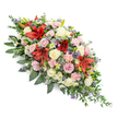 A long, luxurious funeral spray is displayed against a plain white backdrop, giving the impression that it could be gently lifted and placed straight onto a casket for a final journey through Gerrards Cross. The design is lush and full, with an elongated shape that broadens in the centre and tapers gracefully toward each end, suggesting movement and flow rather than rigid formality. Rich red Oriental-style lilies dominate the eye, their large, star-shaped blooms opening outward, each petal velvety and intense in colour, representing deep love and strength in grief. Around them, clusters of soft cream and blush-pink roses, some tightly budded and others fully open, create a sense of tenderness and intimacy, like shared memories whispered at St James's Church or during a quiet gathering at a local chapel. Ruffled pale pink spray carnations run through the arrangement, adding texture and volume, while small, white daisy-like flowers and fine sprays of chrysanthemum blooms fill any empty spaces, ensuring the spray looks abundant from every angle. Cool-toned accents in the form of blue-purple thistles and tiny lavender-blue filler flowers are scattered throughout, bringing a subtle woodland feel that echoes the greenery of nearby common land and gardens. All of this is nestled into a verdant bed of dark green foliage, with broad, polished leaves and softer, trailing greenery that spills out at the edges, particularly near one end, giving the tribute a natural, gently cascading profile. The overall effect is one of composed beauty and heartfelt condolence, a thoughtful floral expression that offers comfort and quiet dignity to families in Gerrards Cross saying their goodbyes.