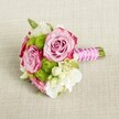 A small, hand-tied posy lies horizontally on a pale, textured linen-style cloth, its colours glowing softly as if caught in late-afternoon light over Gerrards Cross Common. At the heart of the bouquet sit two fully open pink roses and a third rose just behind them, each bloom cupped with layers of velvety petals that move from a gentle blush in the centre to deeper, almost magenta-pink tones at the ruffled outer edges. Their spiralling centres are tight yet inviting, suggesting that the flowers were cut and arranged at peak freshness. Nestled closely around the roses are rounded tufts of lime-green button chrysanthemums, their many tiny florets forming neat, spherical cushions that add playful texture and a modern twist to the classic pink-and-white palette. Between the bolder blooms, clusters of small, creamy white flowers-reminiscent of lisianthus, freesia, or wax flower-thread delicately through the design, their soft petals and tiny star-shaped blossoms adding lightness and gentle movement. Here and there, hints of rich green foliage peek out, giving the bouquet natural depth and a garden-fresh feel, like something gathered from a lovingly tended Chilterns border. The stems are trimmed short and bound tightly into a compact handle, wrapped in a vivid fuchsia satin ribbon that catches the light with a subtle sheen. Over this, a fine strand of white faux pearls winds diagonally, crossing and overlapping as though a bracelet has been draped around the handle, lending a bridal, almost heirloom elegance. Resting on the neutral fabric with its visible weave, the bouquet appears ready to be lifted by a bridesmaid walking into a ceremony in Gerrards Cross, or placed as a delicate centrepiece on a dining table for an intimate celebration, its mood a blend of romance, poise, and quietly joyful anticipation.