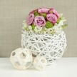 This photograph captures a sophisticated floral centerpiece that feels ideal for a wedding table in Gerrards Cross, blending modern design with soft romance. Anchoring the composition is a round, openwork vase in pure white, built from slim rattan-style strands or twigs that weave and overlap to form a hollow sphere with visible gaps, giving a light, airy impression. The vase stands on a spotless white tabletop, where its sculptural shape is complemented by gentle, even lighting that highlights each curve. Rising from this vessel is a carefully domed bouquet of roses in varying shades of pink and lavender, some blooms fully open with velvety petals unfurling in generous spirals, others slightly tighter with deeper pink edges that add nuance and depth to the colour story. Between the roses, bright green button chrysanthemums offer crisp structure and a fresh, botanical pop of colour, their textured, button-like heads contrasting with the smoothness of the rose petals. Around the lower edge, tiny white filler flowers gather in soft clusters, peeking out and lightly cascading so the bouquet appears relaxed yet still polished. To the left of the vase, resting on the same white surface, sit two decorative balls crafted from pale, natural wood or wicker, each one an open tangle of interlaced strands; the larger sphere sits just beyond the smaller, creating a gentle sense of depth and balance. Behind the arrangement, a blurred backdrop of light beige or tan fabric, reminiscent of linen, adds warmth and subtle texture without competing with the floral colours. The scene suggests a calm, elegant celebration-perhaps a reception near the train station or a private garden wedding off Windsor Road-where thoughtful design and natural beauty come together.