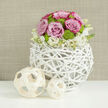 On a pristine white tabletop that subtly reflects every detail, a refined floral centerpiece captures the mood of an elegant wedding reception in Gerrards Cross. The arrangement forms a smooth, dome-shaped bouquet, its structure tight yet inviting, with pastel pink roses opening in generous, velvety spirals. Some of the rose petals deepen towards the edges in a richer blush tone, adding depth and romance to the colour palette. Nestled among them are cheerful lime-green button chrysanthemums, their rounded, compact heads introducing a vivid contrast that feels as fresh as a stroll through East Common after a spring rain. Around the base, small clusters of creamy white blossoms, possibly lisianthus or petite stock, with subtle yellow hearts, seem to flow downwards, softening the line where flowers meet container. These blooms are gathered in an eye-catching spherical vase made from white-painted branches or wicker, woven into an open, cage-like structure that blends modern design with rustic charm. The vase's airy lattice echoes natural textures you might see along the Chilterns, yet its crisp white finish keeps the look contemporary and bridal. To the side, two pale wooden decorative balls, one larger and one smaller, lie on the table, their coiled strips echoing the weave of the vase and completing the visual story without stealing attention. Behind it all, a softly blurred wall in warm taupe tones provides a calm, neutral backdrop. The composition feels serene, joyful, and meticulously crafted - the sort of centrepiece a local florist near the Gerrards Cross station might design for a stylish marquee wedding or chic civil ceremony celebration.