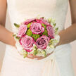 Held gently against the intricate lace of a bridal dress, this wedding bouquet seen in Gerrards Cross captures the intimate pause just before a bride steps out toward the aisle. At its heart, lush lavender-pink roses unfold in full, velvety layers, their colour resting between mauve and dusty blush, like the soft tones of a late-afternoon sky over Gerrards Cross Common. Between these generous petals, vibrant lime-green button chrysanthemums peek through, their rounded, textured heads bringing a lively burst of contrast that keeps the design fresh and contemporary. Around the edges, a ring of creamy-white blooms and slender buds wraps the bouquet in a delicate halo, similar to fine lace edging along a veil or the intricate detail of gowns found in local bridal studios near Packhorse Road. The composition is tightly yet gently gathered, a hand-tied dome that rests securely in the bride's hands, giving a sense of reassurance and calm. Gentle natural light brushes across the flowers, highlighting each curve and contour, while the greenery tucked beneath adds quiet support and structure. In the mind's eye, you can sense a light, romantic fragrance: classic rose sweetness layered with crisp green notes, like walking past freshly tended gardens on Marsham Way. Every flower has been chosen for resilience and beauty, promising to remain radiant from morning photographs in the family home to evening speeches at a nearby country venue, a floral reflection of love, hope, and the start of a new chapter.