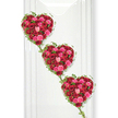 Three rich, rose-filled hearts are arranged one beneath the other, cascading diagonally across a crisp white background in a design that feels both contemporary and deeply sentimental. Each heart is sculpted entirely from tightly clustered roses, the dominant tones a sumptuous crimson red softened by vivid pinks and gentle blush shades that add tenderness and nuance. The petals overlap and curl into one another, creating an almost velvety landscape of colour with subtle changes in height and texture where buds meet full blooms. Encircling these hearts, slim vines of fresh green ivy trace their outlines with graceful precision, the small pointed leaves catching glints of light. These ivy trails extend delicately downwards to touch and bind the hearts together, suggesting an unbroken bond of love, whether between partners, parents and children, or generations of a family. The background is a smooth, bright white surface with the faintest suggestion of vertical lines, much like a refined display wall in a Gerrards Cross chapel or funeral home, allowing the saturated flower colours to stand out vividly. Gentle, even lighting enhances the contrast between the red and pink petals and the cool green foliage, giving the arrangement a polished, almost gallery-like feel. This floral piece would be an especially meaningful tribute for a funeral or memorial in Gerrards Cross, offering a visual expression of affection and remembrance that is both romantic and respectful, as though each individual bloom carries its own quiet message of farewell and love.