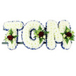 In this photograph, a personalised funeral tribute spells out the name TOM in three bold capital letters, created entirely from fresh white chrysanthemums. The flowers are packed so closely together that they form a smooth, even surface, giving each letter a soft, pillow-like appearance. Around the outer edge of every letter runs a striking royal blue ribbon, pleated into gentle frills to frame the chrysanthemums and enhance the clarity of the name, even when viewed from across a chapel or crematorium aisle in Gerrards Cross. At key points on the design-near the top of the T, along the side and lower curve of the O, and at the upper edge of the M-sit small sprays of accent flowers. These feature velvety red roses as the main blooms, surrounded by dainty white or cream spray roses and slim, trumpet-shaped white flowers that echo the elegance of freesia. The sprays are cushioned by rich green foliage and airy fern fronds, which arch outwards slightly and bring a natural softness and depth to the structured letters. The three floral letters rest together on a slim, dark support bar, indicating a secure backing ideal for resting on a hearse, a trestle, or beside the coffin during a memorial service in local churches around Station Road or West Common. Set against a bright white studio-style background, the arrangement's clean lines and carefully chosen colours convey purity, peace, love, and remembrance, creating a respectful floral farewell for someone named Tom from his Gerrards Cross community.