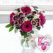 This image shows a joyful floral gift set that feels perfectly suited to a sunny home in Gerrards Cross, perhaps just delivered to a doorstep off Oxford Road. In the centre, a rounded bouquet brims with fresh blooms arranged in a clear glass vase. The vase is generously filled with water, and the stems inside are neatly trimmed and splayed, showing the care of an experienced florist. Around the neck of the vase, natural-coloured twine is tied in a relaxed bow, from which a small, white, rectangular tag hangs, featuring a simple pink rose motif and green script, giving the impression of hand-selected flowers. The bouquet's focal points are large gerbera daisies in a glowing magenta or deep fuchsia tone, each with a dark, velvety centre that creates striking contrast. Clustered between them are numerous two-toned chrysanthemums: their petals begin as deep purple near the centre and fade to clean white tips, circling bright yellow hearts that add a sunny, playful note. Soft, creamy white roses are nestled throughout, some still in bud and others gently unfolding, lending a graceful, romantic touch. Sprays of fine baby's breath weave through the arrangement like delicate lace, filling gaps and softening the transitions between colours. Fresh, glossy greenery frames the blossoms and helps create a full, rounded silhouette, reminiscent of a garden border in bloom. To the lower right of the vase, a shiny heart-shaped foil balloon edged in silver adds a fun, celebratory accent. Its white centre is decorated with bold, multi-coloured letters spelling out Love n' Hugs 24-7, completing the theme of constant affection. The bouquet and balloon rest on a pale, spotless surface, while the softly blurred, bright background enhances the sense of a light-filled, uplifting flower delivery in Gerrards Cross.