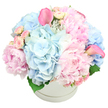 A sumptuous round hatbox filled with pastel blooms is shown against a bright white backdrop, every detail carefully composed to highlight the flowers' soft elegance. The container is a pristine white, circular box with a delicate ridge around its edge, giving a refined gift-like quality that would sit beautifully on a console table in a Gerrards Cross hallway near Fulmer Road. Overflowing from the top, clusters of light blue hydrangeas form a cool, pillowy base, their many tiny blossoms merging into generous, cloudlike domes. Nestled among them, large pink peonies open in luxurious layers, their ruffled petals creating a romantic, almost whipped-cream texture that draws the eye to the centre of the design. Smaller, blush spray roses are dotted throughout, some in tight buds with smooth outer petals, others beginning to unfurl, introducing intricate detail and a sense of unfolding stories. Two striking pink calla lilies rise from within the arrangement-one angled towards the upper left, the other to the right-bringing sleek, contemporary curves that contrast beautifully with the softer, rounder petals. A few deep green leaves sit low around the base of the blooms, just visible above the rim of the hatbox and adding subtle depth and structure. The flowers are tightly massed into a rounded dome that gently spills over the edge, giving an impression of generous, carefully curated abundance. The mood is tender, joyful, and soothing at once, making this arrangement an ideal choice for a new baby celebration, a thoughtful get-well surprise, an elegant Mother's Day gift, or a refined sympathy gesture delivered locally across Gerrards Cross.