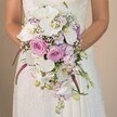 A bride, captured from the waist up in the heart of Gerrards Cross, delicately cradles a show-stopping cascading bouquet, her white lace gown shimmering softly against a pale backdrop. This exquisitely styled wedding arrangement draws immediate focus with its luxurious layering of pure white Phalaenopsis orchids, their wide petals and gold-flecked centers gleaming in the gentle light reminiscent of the refined interiors found along Packhorse Road. Surrounding the orchids, lush blush pink and lavender roses unfold-some in tight spirals, others blooming wide-adding classic romance and a fresh floral scent that brings to mind spring gardens across Gerrards Cross. Wisps of baby's breath cascade between the larger flowers, creating a feathery, cloud-like dimension, while strands of vivid magenta Amaranthus extend downward, adding movement and a touch of dramatic flair. Intertwined lavender blooms, likely sweet pea or stock, enrich the palette, offset by the naturally trailing green foliage and slender unopened buds, each detail skillfully arranged to reflect both modern taste and timeless elegance. The entire bouquet glows with the blended colours of white, pink, and purple, anchored by flecks of green, making it an ideal choice for a luxury wedding or to inspire couples planning nuptials in and around Gerrards Cross. This floral composition not only captures the essence of bridal sophistication but also celebrates the artistry and fresh creativity found with local Gerrards Cross florists.