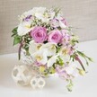A beautifully composed floral arrangement sits in a white wicker basket, overflowing with soft pink and white blooms that feel perfectly suited to a wedding table in Gerrards Cross. The eye is drawn first to the fully opened pastel pink roses, their petals layered in tight spirals that radiate outwards and give a sense of warmth and romance. Surrounding them, white Phalaenopsis orchids extend their broad, almost porcelain-like petals, each blossom showcasing a luminous yellow and subtle orange centre that adds an exotic note. Nestled between these star flowers, white Lisianthus blooms contribute a more relaxed, ruffled texture, with some petals edged in a gentle lavender-purple hue that introduces a touch of whimsy. Pale pink stock flowers rise and then drift softly over the edges of the basket, their slender stems and clustered petals adding both height and movement, as if the bouquet is gently breathing. Scattered throughout, delicate clouds of white baby's breath create light, airy pockets that prevent the arrangement from feeling heavy, while glossy green leaves and a few trailing buds peek from beneath, hinting at new growth and freshness. The basket itself is rounded and neatly woven in white, its rustic charm echoed by two decorative woven spheres placed on the crisp white surface beside it, like small natural sculptures. Behind, a neutral, beige-toned fabric backdrop-suggestive of linen-adds soft texture without distracting from the flowers. The whole scene feels calm, romantic and carefully curated, the kind of centrepiece you might find at an intimate celebration near Gerrards Cross station or in a private dining room off Packhorse Road, ready to be enjoyed by guests gathered around the table.