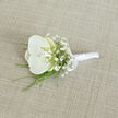 Displayed diagonally on a softly woven, light tan fabric that resembles an elegant suit swatch, this white orchid boutonnière captures the understated refinement often chosen by couples marrying in and around Gerrards Cross. At its heart is a single, immaculate orchid bloom, its rounded petals satin-smooth and luminous, radiating a clean white that gradually warms toward the centre where a whisper of pale yellow and cream glows in the throat. This gentle shift in tone creates a feeling of depth and draws the eye inward, much like a focal point in a carefully composed bridal bouquet. Surrounding the orchid, delicate sprays of baby's breath scatter tiny white blossoms in all directions, forming a fine, cloud-like halo that softens the stronger lines of the main flower. From beneath the bloom, slim blades of bright green foliage - likely decorative grasses or fine fern fronds - extend in graceful arcs, offering freshness and a crisp contrast, reminiscent of the greenery lining the quiet lanes near West Common and the leafy gardens around the village centre. The stems of the orchid and filler flowers are expertly gathered and bound, wrapped in a smooth, gleaming white satin ribbon that creates a sleek, cylindrical handle, practical for pinning yet beautifully finished. A small, perfectly tied satin bow sits just under the base of the flowers, giving a hint of classical charm without overpowering the design. The fabric beneath has a subtle, visible weave that adds delicate texture, preventing the background from competing with the floral details while still grounding the image in a tactile reality. Soft, even lighting illuminates every curve of petal and thread of ribbon, evoking calm sophistication and the poised stillness that precedes a ceremony, as if this boutonnière is waiting on a dressing table before being carried to St. James's or a local Gerrards Cross venue. The overall mood is one of quiet elegance and heartfelt intention, perfect for a groom or honoured guest.