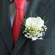 This image shows a finely tailored charcoal jacket in Gerrards Cross, its breast pocket transformed into a small floral showcase by a graceful white orchid boutonnière. The orchid sits centrally, its broad, smooth petals unfolding with quiet elegance, their creamy surface catching gentle light in a soft, almost velvety glow. At its heart, a hint of pale gold peeks through, adding depth and a sense of hidden emotion. A delicate drift of baby's breath surrounds the bloom, each tiny white flower like a suspended droplet of mist, softening the edges and lending an ethereal, romantic air. Threads of fresh green foliage weave through and around the cluster, adding movement and a subtle woodland energy, reminiscent of the leafy lanes leading out of Gerrards Cross towards the Chilterns. At the base, a slim white ribbon is tied in a clean, unfussy bow, its satin folds pressed neatly, suggesting the careful touch of an experienced florist. Against the matte charcoal fabric of the suit and the bold, jewel-like red of the silk tie, the boutonnière stands out with refined brightness, as though illuminated from within. It feels like a piece chosen for a wedding or formal celebration in one of the elegant local venues, capturing the promise of the day in a single, serene flower. The overall effect is poised and romantic, a quiet statement of love and ceremony worn close to the heart.