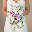 The image captures a refined, waist-level view of a bride holding a cascading wedding bouquet, her white strapless gown forming a clean, luminous backdrop for the flowers. The arrangement is composed in a dreamy palette of white, pink, and soft lavender, with the flowers gathered tightly at the top and then flowing down in an elegant teardrop trail. At the heart of the bouquet are breathtaking white Phalaenopsis orchids, their large, smooth petals almost porcelain-like, each bloom revealing a delicate yellow-orange centre that adds warmth to the cool tones. Nestled among them are classic pink roses and slightly ruffled, garden-style roses in dusty lavender-pink, their many petals giving the bouquet a plush, romantic fullness. Around these focal blooms, you can see white lisianthus with subtle purple-edged petals, their slender buds curling gently, and fine stems of baby's breath scattered like a soft, white haze. Pale lavender stock adds vertical interest and a hint of country-garden charm, while hidden greenery supports the cascade without distracting from the pastel harmony. The background is softly blurred and beige, ensuring every detail of the bouquet stands out clearly, almost as if you could reach in and feel the contrasting textures. This style of bridal bouquet would feel completely at home in a Gerrards Cross wedding, whether carried down the aisle at a church off Packhorse Road or in a nearby country venue, and it reflects the kind of bespoke, romantic floral craftsmanship couples often seek from experienced local florists in the area.