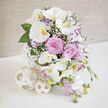 A refined floral arrangement sits poised on a clean white surface, its colours and textures evoking the quiet elegance of a wedding breakfast in a Gerrards Cross townhouse. The central focus is a cluster of pristine white Phalaenopsis orchids, their broad petals smooth and luminous, curving outward like butterfly wings. Each orchid face is detailed with a golden-yellow and soft orange centre, drawing the eye in with an almost glowing intensity. Around these exotic blooms, generously opened roses in a muted lavender-pink shade spiral gently outward, their layers of velvety petals creating a sense of depth and romance. Threaded throughout, tiny white baby's breath forms a delicate mist, softening the transitions between the larger flowers and giving the bouquet a dreamy, cloud-like quality. Slender stems of pale purple lisianthus, some tinted with deeper violet along the petal edges, extend the arrangement outward, while cascades of petite lavender-pink flowers, like fragrant stock or sweet peas, drift downward in graceful streams. Lush green leaves, from smooth, broad foliage to tender new buds, bring structure and a fresh garden feel, reminiscent of the greenery around East Common. All of the blooms are gathered into a striking, contemporary white vase made from narrow interwoven strips forming an open, spherical lattice. At the base, two coordinating decorative orbs crafted from similar light material rest nearby, echoing the vase's airy geometry. Behind the display, a softly textured beige backdrop, bathed in bright yet gentle natural light, enhances every pastel tone and glossy leaf. Crafted with the care of a seasoned local florist, this piece feels perfectly suited to wedding receptions, anniversaries, or intimate celebrations at home, and would translate beautifully to same-day delivery across Gerrards Cross.