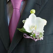 Against the backdrop of a deep charcoal suit, a pristine white orchid boutonniere becomes the gentle highlight of a formal moment, much like a calm pause amid the bustle of Gerrards Cross station at rush hour. The orchid's petals are wide and velvety, their ivory tone almost glowing, with a soft translucence that hints at delicacy without fragility. Nestled around this central bloom are slender, silvery-green leaves and sprigs of lilac and lavender, each tiny flowerhead adding nuance in texture and colour, like the subtle shifts of light over the gardens near West Common. Wisps of white gypsophila tuck in between, lending an airy softness that keeps the composition light and modern. A narrow violet ribbon, neatly wrapped and tied, hugs the stems and echoes the purple-pink tie worn beneath the jacket, creating a harmonious palette of white, green, and gentle mauve. The overall effect is both polished and romantic, ideal for a wedding ceremony at a local church or a reception in one of the nearby country venues. The boutonniere sits precisely at the lapel, suggesting care in every detail, as though it has just been pinned on moments before stepping into photographs on Packhorse Road. There is a subtle sense of fragrance-a mix of fresh greenery and powdery florals-that mirrors the soft, hopeful mood of the day. This understated arrangement speaks of elegance and personal expression, capturing the fleeting beauty of a promise made and celebrated close to home.