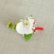 A refined boutonnière rests on a pale, linen-like fabric that has a soft, woven texture, the gentle beige tone creating a calm, natural stage for the design. At the centre sits a large, pristine white orchid, its petals broad and satin-smooth, curving outwards with effortless grace. The inner throat of the orchid glows subtly with shades of creamy yellow and tiny orange flecks, drawing the eye inward and adding depth to the otherwise pure white bloom. Around this focal flower, sprigs of dainty white baby's breath cluster like miniature clouds, bringing a sense of lightness and romance that feels perfect for a wedding ceremony or formal celebration in Gerrards Cross. Just to the left, two compact, bright green buds-suggestive of green button chrysanthemums or a similar mum-offer a playful, textured contrast, their slightly spiky forms giving energy and movement to the composition. A single glossy green leaf, likely Ruscus, emerges from beneath the orchid, adding structure and a touch of woodland freshness, as though the flowers were gathered near one of the leafy paths towards Gerrards Cross Common. The stems are enveloped in a vivid red satin ribbon, wrapped cleanly and tied into a neat little bow on the right side, its polished sheen catching hints of light and symbolising passion, joy, and celebration. This precise combination of crisp white, verdant green, and rich red creates a timeless accessory suited for a groom's suit, a stylish prom outfit, or any special occasion where elegant understatement is cherished. You can almost imagine this piece being pinned just before stepping into St James's Church or a local reception venue, a final, meaningful flourish crafted by an experienced florist who understands how small details can transform a moment.