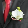 A close-up photograph reveals an elegant wedding boutonniere pinned with precision to the lapel of a dark charcoal grey suit jacket, its fine wool fabric showing a subtle woven texture in the soft, indoor light. Beside it, a bold, solid red satin tie lies against a light grey and white small-checked dress shirt, the smooth silk catching gentle highlights and echoing the romance of a wedding in Gerrards Cross. At the heart of the boutonniere, a single, large white Phalaenopsis orchid opens gracefully, its broad, velvety petals curving outward to frame a distinct yellow and white lip at the centre. Clusters of tiny white baby's breath surround the orchid like a delicate cloud, each miniature bloom airy and precise, while a few rich green leaves fan out behind, providing a lush, natural frame. Two neat, round lime-green button chrysanthemum-style blooms nestle just above the orchid, their bright colour and textured surfaces adding a playful, contemporary touch that would feel at home at a ceremony near East Common or St. James's Church. The stems of the flowers are wrapped tightly in a deep red satin ribbon that subtly matches the groom's tie, its narrow band creating a polished finish at the base of the design. The boutonniere sits just above a neatly stitched jacket pocket, the composition highlighting the careful craftsmanship of a professional florist from the Gerrards Cross area and the quiet anticipation of a special day. The overall mood is refined, romantic, and thoughtfully modern, as if captured moments before the groom steps out for photographs in one of the town's leafy streets.