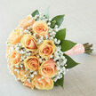 There is a portrait-like tenderness to this hand-tied bouquet: apricot-peach roses arranged in a compact, rounded cluster so their creamy petals overlap like silk folds, each bloom chosen for symmetry and softness. Tiny white gypsophila drifts between the roses, a delicate mist of blossoms that adds air and breathes lightness into the design; the white points contrast the peach warmth and draw the eye inward. Spear-like, glossy deep-green leaves are artfully interspersed, giving the bouquet structure and a fresh, emerald depth that frames the roses without stealing the spotlight. The stems are finished with a textured peach ribbon studded with pearls - a careful, tactile detail that lends a refined, personal touch to the bouquet's base. Bathed in soft daylight, the arrangement glows subtly, the pastel tones and the sheen on the petals highlighted by gentle diffuse lighting; the neutral studio setting keeps attention on the floral shapes and fine textures. The scent is understated and clean: green, leafy notes of freshly cut stems blending with the soft, honeyed breath of roses and the faint powdery sweetness of baby's breath. This bouquet from a local hands-on studio is exactly the sort of arrangement we deliver around Gerrards Cross station and the High Street for weddings, vow renewals, or intimate anniversary gatherings; at Flower Delivery Gerrards Cross we take care to present each bouquet so it reads as both an emblem of affection and an heirloom to be held close.