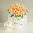 This hand-arranged bouquet marries contemporary sculpture with classic romance: a dome of honeyed-peach roses rests above a round, white woven basket whose open lattice gives the entire piece a sense of buoyancy. Each rose unfurls in soft spirals, the petals shifting from pale cream to warm apricot at their centers, inviting slow admiration and a faint, sweet scent that drifts gently through the air. Sprays of delicate white gypsophila weave through the roses like a lace veil, their tiny blossoms scattering light and creating a froth of texture that contrasts wonderfully with the smoother rose petals. Fresh green foliage frames the dome, the leaves' cool, satiny surfaces adding depth and a hint of herbal freshness. The spherical rattan base is both decorative and architectural, its organic lines echoing trends in modern interiors while remaining soft enough for a romantic setting-ideal for a Gerrards Cross wedding, a thoughtful gift from friends returning from a walk around the town green, or a refined centrepiece for a celebration on Station Road. As a local florist I pay attention to balance and scent; this arrangement feels like a morning in spring, calm and hopeful. Available for local delivery around Gerrards Cross, it's crafted to bring understated elegance and the quiet joy of new beginnings to any occasion handled with care by Flower Delivery Gerrards Cross.