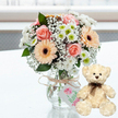 A softly romantic arrangement is displayed on a clean white surface, evoking the relaxed elegance of an anniversary celebration in Gerrards Cross. At the heart of the scene, a glass jar vase holds a compact, dome-shaped bouquet of fresh flowers. Pale pink roses with velvety petals are nestled beside peach gerbera daisies, their dark centres adding depth against the pastel tones. Around them, clusters of crisp white chrysanthemums with hints of lime green at their centres and delicate sprays of white baby's breath create a light, cloudlike halo, balanced by discreet touches of dark green foliage. The neck of the vase is wrapped in rustic natural twine, the bow carefully tied, and from it hangs a small pale gift tag printed with a soft pink floral emblem and fine green script, reinforcing the sense of something lovingly assembled by hand. To the right, a small cream teddy bear sits upright, its body turned slightly towards the bouquet as though sharing in the sentiment. The bear's fur is plush and textured, its dark bead eyes and brown stitched nose giving it a gentle, reassuring expression, while a narrow brown satin ribbon is knotted neatly at its throat. The polished white surface provides a faint reflection, enhancing the feeling of freshness and light, and the gently blurred background hints at a bright interior, perhaps near the station or close to the shops in Gerrards Cross village. Together, the pastel flowers and cuddly teddy bear create a scene that speaks of tenderness, appreciation, and enduring companionship, ideal for marking wedding anniversaries, relationship milestones, or heartfelt gestures of thanks.