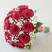 A strikingly elegant bouquet takes center stage, reflecting the charm and intimacy of a Gerrards Cross celebration. Against a pale, softly textured surface, countless deep red roses in full, plush bloom gather closely together, their rich, velvety petals forming a dramatic dome that visually whispers of passion and devotion. Interlaced among these vibrant blooms are delicate tufts of snowy-white baby's breath, offering a luminous contrast and airy softness, much like the gentle light that filters through the leafy lanes off Fulmer Road. Subtle hints of rich green foliage peek from between flowers, providing natural depth and balance. The base features a handle wearer could easily grasp, smartly sheathed in a wide scarlet satin ribbon accented by clean white stripes and a shimmering strand of pearl beads spiraling gracefully, signaling readiness for a walk down the aisle. Trimmed green stems show through at the base, a tangible nod to freshness and local craftsmanship. Illuminated by even, natural daylight common in Buckinghamshire's mornings, the bouquet's vivid hues and intricate detailing catch every eye, evoking emotions of love, festivity, and intimacy-ideal for weddings, vow renewals, or romantic gifts delivered to homes throughout Gerrards Cross. This arrangement beautifully fuses tradition and artistry, its meaning deepened by the community spirit woven through every petal.