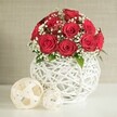 A romantic red rose arrangement is the undisputed focal point of this image, perfectly suited to a wedding table or special celebration near Gerrards Cross. The bouquet forms a generous dome of deep crimson roses, each flower in full, luxurious bloom so that their velvety petals fold over one another in intricate spirals. The roses are densely packed, creating a sense of fullness and abundance, while delicate clusters of white baby's breath weave between them, adding a lace-like lightness that feels almost bridal. Flecks of fresh green foliage appear sparingly at the edges and between some blossoms, their subtle presence enhancing the natural beauty without distracting from the rich red-and-white contrast. The arrangement is set in a unique, bulbous white vase whose entire surface is made up of interlocking strands, as though fine white twigs or rattan have been carefully shaped into a hollow sphere. This open, woven design allows glimpses of shadow and light to play across the structure, giving it a sculptural, handcrafted quality. The vase rests on a smooth, white tabletop, which reflects a little of the soft, ambient light and keeps the scene clean and uncluttered. To the left of the vase sit two decorative woven spheres in a creamy, off-white shade, one large and one small, their loose lattice pattern echoing the design of the vase and tying the composition together in a harmonious way. Behind everything, a gently blurred, neutral-coloured wall creates a calm backdrop that lets the vibrant roses remain the star. The effect is of quiet luxury and heartfelt emotion, evoking the atmosphere of an intimate wedding reception or anniversary dinner styled with care by a local Gerrards Cross florist.