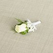 A classic single white rose boutonniere lies diagonally across a softly textured, light beige fabric that resembles finely woven linen, the kind you might see on an elegant table in a Gerrards Cross townhouse. At the heart of the image is a creamy white rosebud, its velvety petals still gently furled, suggesting a bloom cut at perfect freshness just hours before a wedding at the local church or a ceremony in one of the leafy avenues off Packhorse Road. Around the rose, clusters of delicate white baby's breath form tiny clouds of blossom, each minuscule flower adding a lace-like halo that softens the overall shape and gives a feeling of quiet romance. Slim, deep green leaves and a few more feathery sprigs of foliage peek from behind the bloom, their rich colour and subtle sheen offering a natural contrast to the rose's ivory tones and the neutral backdrop. The stems are neatly bound in a pristine white satin ribbon, wrapped with the steady, practiced hands of an experienced florist, then finished with a small, perfectly tied bow that sits just under the flower head, ready to be pinned to a lapel. Soft, even lighting falls across the boutonniere, picking up the gentle curves of each petal, the fine grain of the fabric beneath, and the faint shadows where greenery overlaps. The overall mood is serene and sophisticated, evoking the anticipation of a groom or groomsman dressing for a special day in Gerrards Cross, perhaps before photographs in nearby East Common or a reception in a local hotel, where this understated white rose will quietly symbolise purity, commitment, and timeless style.