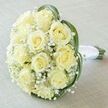 There is a nearly dreamlike hush to this hand-tied bridal bouquet-an arrangement of approximately fifteen to twenty creamy yellow roses whose gently spiraled petals form a compact, luscious dome of pale ivory and warm cream. The roses glow with a soft inner light, their velvety faces edged with the faintest touch of vanilla, while delicate sprays of Gypsophila (baby's breath) drift between them like small white clouds, giving the bouquet a featherlight, ethereal feeling. Slim, vibrant blades of green grass-likely lily or bear grass-arc gracefully around the edge, creating a modern, natural frame that suggests a slow, elegant motion. At the base, the green stems are gathered and wrapped in pristine white satin ribbon, tied with the careful hand of an experienced florist so the handle sits cool and secure in the hand. The bouquet rests on a pale, textured linen backdrop in light taupe or off-white; soft, diffused lighting from a morning window reveals subtle shifts in the roses' creamy tones and gently highlights the airy Gypsophila. As a florist with Flower Delivery Gerrards Cross who often arranges flowers for weddings near the station and for ceremonies by Denham Country Park, I imagine this bouquet carried down a chapel aisle or presented at a small, intimate celebration. It communicates purity, quiet joy, and a refined simplicity-an ideal choice for brides who want classic romance with a contemporary, local touch and for anyone in Gerrards Cross seeking a bouquet that feels both familiar and exceptionally crafted.
