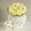 An elegant wedding table arrangement is presented against a softly blurred, neutral backdrop, as if ready to be placed on a dressed table in a Gerrards Cross reception room. At the heart of the composition is a generous, dome-shaped cluster of pale yellow roses, tightly massed so that their velvety petals seem to form one continuous cushion of soft colour. Each rose is flawless, their tones shifting subtly from buttery cream at the outer edges to a richer pastel yellow toward the centres, giving the bouquet depth and a gentle inner glow. Threaded delicately between these blooms are fine sprays of white baby's breath, their many tiny blossoms forming a light, cloud-like mist that softens the rose contours and adds texture without disturbing the calm palette. A slim circlet of rich green foliage sits neatly around the base, the smooth leaves creating a polished collar that emphasises the careful craftsmanship of the florist. The flowers emerge from a distinctive white woven vase, whose rounded form and open, basket-style construction suggest modern design inspired by natural materials. Light seeps through its crisscrossed strands and falls onto the glossy white tabletop, creating fleeting shadows that echo the weave. To one side, two decorative spheres made from pale, rattan-like strips continue the motif, their airy geometric patterns providing balance and interest without competing with the flowers. The background, a soft beige with a hint of texture, remains gently out of focus, allowing the fresh yellows, whites, and greens to stand forward. The overall impression is one of refined simplicity and quiet joy, an arrangement perfectly at home on a bridal table or intimate celebration near the heart of Gerrards Cross.