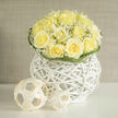 This bouquet reads as an understated celebration in ivory and soft lemon: a neat dome of creamy yellow roses embraced by airy sprays of baby's breath, all arranged as a single sculptural statement within a white rattan orb. The roses are gathered closely so that their ruffled centers open like small, contented sighs, while the gypsophila threads through the petals like scattered confetti, catching light and adding a cloud-like softness. The rattan vessel acts both as container and architectural base - a tactile, modern pedestal whose whitened weave echoes the spherical woven decorations placed beside it. Those minimalist spheres provide a playful counterpoint, their natural fibres and geometric presence amplifying the arrangement's contemporary sensibility without stealing its quiet voice. There is a sensory warmth to the scene: a hint of meadow-sweet and fresh-cut stems underscored by a subtle, grounding earth note, and the petals themselves promise a velvety, almost linen-like touch. I imagine this piece arriving at a calm morning gathering on Packhorse Road or brightening a reception table at a small celebration in nearby Chalfont St Peter, where neighbours appreciate restrained elegance. As a local florist I choose these materials to speak of thoughtful restraint and optimism - suitable for congratulatory messages, gentle encouragements, or as a refined accent at an intimate event. Flower Delivery Gerrards Cross has arranged many such compositions for residents who favour both simplicity and sculptural beauty in their floral choices.