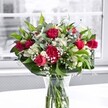 A lush, romantic bouquet of red roses and matching crimson carnations stands proudly in a clear glass vase on a bright white tabletop, bathed in gentle daylight from a large window that looks out over a softly blurred Gerrards Cross street scene. The roses vary from tight, promising buds to fully unfurled blooms with velvety petals that curl back in layered spirals, while the carnations add ruffled depth, their frilled edges catching the light and giving the arrangement a plush, almost tactile richness. Threaded between the deep reds are pristine white alstroemeria, some still in slender green-tipped bud, others beginning to open into trumpet-shaped flowers with delicate, speckled throats, suggesting days of unfolding beauty. Tiny white gypsophila, or baby's breath, drifts like a light mist through the bouquet, softening the stronger colours and giving the composition an airy, cloudlike halo. Around the base, broad, glossy green leaves form a structured collar, their surfaces smooth and reflective, while finer, lighter greenery arches and weaves between the blooms, adding movement and a sense of garden freshness. Through the crystal-clear vase, the neatly trimmed stems are visible, standing in clean water that glows faintly where it catches the reflection of the white table. The background remains desaturated and out of focus, so all attention rests on the vivid reds, crisp whites, and vibrant greens of the fresh flowers. The scene feels like a quiet celebration-a bouquet suited to an anniversary dinner on a Gerrards Cross High Street apartment windowsill, a thoughtful thank-you delivered across town, or a simple gesture to brighten a home near East Common with colour, fragrance, and the calm joy of fresh flowers.