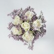 An exquisite overhead photograph shows a compact hand-tied bouquet resting on a pale, stone-like surface, its circular shape perfectly framed from above. At the heart of the arrangement sit six creamy white roses, each bloom fully formed yet still delicately furled, with velvety petals spiralling inward to create a sense of depth and quiet luxury. Hints of pale green can be seen at the base of some outer petals and along the sepals, lending a fresh, almost dewy quality that feels like an early morning walk through the calm streets of Gerrards Cross before the shops open. Surrounding these pristine roses is a soft halo of tiny lavender and muted lilac limonium, or statice, creating a feathery mist of colour that gently cushions each rose. The petite star-shaped blossoms are densely clustered, but light in appearance, like a cloud of wildflowers gathered from the edges of the woodland near West Common. The cool, even lighting casts no harsh shadows, instead allowing the ivory tones of the roses to glow subtly against the purple haze around them. The smooth background in off-white or very light grey keeps the eye focused entirely on the flowers, emphasising their texture, from the plush rose petals to the papery fineness of the filler blooms. The overall mood is one of serenity, tenderness, and understated refinement, perfectly suited to a sympathy bouquet, a thoughtful get-well gesture, or a peaceful bridal posy for an intimate ceremony in one of Gerrards Cross's local churches. The image quietly conveys care and craftsmanship, reflecting the kind of elegant, hand-crafted design a local florist might prepare for same-day delivery to a nearby home or small gathering.