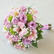 Resting softly on a pale, neutral cloth, this contemporary bridal bouquet glows with harmonious pink, lilac, white, and green tones, echoing the refined aesthetics often admired in Gerrards Cross homes. Plump, full lavender roses stand as the focal point, their spiral petals capturing gentle sunlight as it might filter through the mature trees along Marsham Lane. Interwoven are lively clusters of lime green button mums, their rounded forms providing exuberant contrast and a modern twist to the romantic pastels. Soft pink Alstroemeria lilies, adorned with delicate maroon veining and golden highlights, sweep outward with graceful movement reminiscent of Gerrards Cross's spring garden walks. Generous clouds of baby's breath mingle between blossoms, the tiny white blooms introducing an air of lightness and effortless charm. Dark, glossy leaves peep from beneath, hinting at the bouquet's fresh-picked vibrancy and the lushness of local greenery. Carefully gathered and wrapped in a shimmering rose-pink satin ribbon, finished with a tender strand of pearls, each stem is secured for easy handling and elegant presentation-befitting both weddings in the heart of Gerrards Cross and heartfelt floral gifts for milestone celebrations. This sophisticated, expertly crafted bouquet exemplifies the balance of tradition and modernity found in Gerrards Cross floristry, its textures and colours layered with intention and artistry.