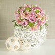 Bathed in gentle studio light, a handcrafted floral centerpiece sits invitingly on a white surface, embodying the relaxed yet refined charm of Gerrards Cross. Arranged with an artist's touch, the bouquet overflows with soft pastel pink and lavender roses, plush and opulent, interspersed with striped pink alstroemeria that add both movement and gentle contrast. Cheerful lime green button chrysanthemums punctuate the design with bursts of freshness, while airy clusters of white baby's breath create a feathery, ethereal halo-reminiscent of early blooms in Chalfont Park. The flowers are cradled in an eye-catching white vase featuring an intricate woven lattice, its organic shape and open pattern balancing contemporary design with a rustic nod to local artisan traditions. Flanking the arrangement, two woven spheres fashioned from light, natural fibers echo the vase's texture; the larger nestles closer for stability, while the smaller subtly advances, adding playful asymmetry and textural harmony. All elements come together against a softly textured beige background, lending the composition a sense of peace and understated luxury. Perfect for wedding tables or sophisticated home gatherings in Gerrards Cross, this arrangement showcases the imaginative craftsmanship and attention to natural beauty characteristic of the area's finest florists.