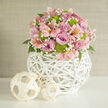 This close-up image captures a carefully crafted floral centrepiece that would sit beautifully on a wedding table in one of Gerrards Cross's elegant venues near the Common. The arrangement forms a full, dome-like bouquet, where every space is thoughtfully filled yet nothing feels crowded. Soft lavender and blush pink roses dominate the design, their petals spiralling outward in perfect, velvety layers that suggest romance and gentle celebration. Between them, pale pink alstroemeria - the familiar Peruvian lilies - stretch delicate petals marked with fine speckles and stripes, adding a sense of movement and playfulness. Bright lime green button chrysanthemums punctuate the pastels with their crisp, modern energy, their compact, rounded blooms mirroring the shape of the overall design and giving a fresh, almost zesty accent, like the fresh greenery around Bulstrode Park. Wisps of white baby's breath weave around the larger flowers, creating a soft haze that diffuses the light and emphasizes the bouquet's softness. Deep green foliage peeks from underneath, offering contrast and framing the pastel flowers without overpowering them. The whole arrangement is nestled in a striking white woven globe-shaped vase, crafted from intricately interlaced wicker or rattan, its open lattice giving a feeling of lightness and air. Next to it on the smooth white surface lie two smaller woven decorative spheres in a paler, natural wood shade, echoing the texture of the vase and adding a curated, interior-styling touch. The neutral, off-white backdrop ensures every tone and texture stands out clearly. The mood is refined yet inviting, ideal for wedding flowers or stylish home décor, and reflects the kind of thoughtful design a local Gerrards Cross florist might create for same-day delivery.