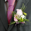 This image shows a refined wedding boutonnière, created with the care and balance you might expect from a seasoned florist working in Gerrards Cross. At its core sits a single pale pink rose, its petals just beginning to unfurl in a gentle spiral, giving a sense of soft movement and budding romance. Clustered around the rose, airy white gypsophila forms a delicate surround, each tiny blossom like a star in a light, floral constellation. Nestled alongside, a bright green button chrysanthemum offers a standout focal point, its rounded shape and fresh lime tone adding a contemporary twist to the more traditional rose. Slender fronds of greenery and a longer blade of foliage stretch slightly beyond the flowers, giving the boutonnière a slim, elegant silhouette that would complement a tailored suit at a registry office ceremony or a celebration near West Common. The stems are neatly wrapped in a muted mauve ribbon, tied with precision so the ends fall gracefully, adding just a hint of colour and softness against the jacket. Though you can't smell it in the photograph, the gentle scent of rose would likely linger as the wearer greets guests or walks through one of the leafy streets near Station Road. Every element is composed to feel balanced and intentional, yet not overdone, making this boutonnière a subtle but meaningful accent for weddings, proms, or formal events where small details express big emotions.