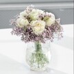 A softly lit close-up of a graceful white and lavender bouquet shows creamy white roses opening in gentle spirals, each petal edged with a faint glow of pale yellow at the centre, as if just unfurling in the morning light. Around them drifts a cloud of tiny lavender and lilac statice flowers, their papery clusters forming a mist-like halo that adds both texture and airy volume to the design. The stems gather neatly in a clear glass vase with a distinctive crackle, or ice-effect, finish; the textured surface catches every glint of light and scatters it in fine reflections across the smooth white tabletop beneath. Through the transparent water, the strong green stems are clearly visible, creating a simple, natural structure that anchors the arrangement. Behind the bouquet, the background falls away into a soft blur of pale grey and gentle light, making the flowers appear even more serene and luminous. The mood is calm and contemplative, like a quiet corner of a home near Gerrards Cross Common or a sunlit windowsill off Packhorse Road. This is the kind of arrangement that could rest beside a bed in a Gerrards Cross townhouse, soothe the atmosphere in a small clinic reception, or offer a tender message of support. Every detail-from the choice of white roses to the feathery veil of statice-suggests a florist's careful hand and an intention to bring quiet comfort and understated elegance into an everyday Buckinghamshire setting.