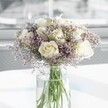 A full, rounded bouquet of creamy white roses and soft lavender statice stands at the centre of the image, arranged in a tall, clear glass cylinder vase on a bright white tabletop. Around a dozen white roses form the heart of the design, each bloom at that perfect stage of opening where the petals gently unfurl, revealing spiralled centres with a subtle ivory glow. The petals look velvety and thick, catching the soft natural light that appears to stream in from a large nearby window, perhaps like those in the airy homes off Packhorse Road in Gerrards Cross. Threaded carefully between the roses are feathery clusters of tiny lilac-purple statice, their papery flowers creating an almost cloud-like halo that blurs the edges of the bouquet. The stems are clearly visible through the glass, a bundle of fresh, straight green lines submerged in clear water that glints faintly in the daylight. The background is softly out of focus, hinting at a calm indoor space rather than drawing attention away from the flowers themselves. The overall mood feels serene and polished, like a bouquet chosen to bring quiet comfort to a memorial service, to brighten a minimalist living room near West Common, or to sit gracefully on a bedside table after a meaningful celebration. There is no ribbon or extra foliage, just the interplay of white and lavender, rose and statice, classic and delicate. The arrangement communicates a gentle sense of hope and remembrance, the sort of thoughtfully balanced design a local florist might recommend when you want your flowers to speak with subtlety rather than flourish.
