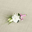 Resting on a subtly textured, pale woven cloth, this boutonniere feels like a whispered moment of romance captured in flowers, the sort of keepsake you might wear for a ceremony in a Gerrards Cross church or at a celebration near the Memorial Centre. The centrepiece is a fully open white lisianthus bloom, its petals luxuriously layered and softly rippled, creating a cushion of creamy white that seems almost weightless. Beside it, two lisianthus buds lean closely in, still partially closed, their gently rounded forms symbolising the beginning of a story yet to unfold. Fine sprigs of baby's breath weave lightly through the composition, their tiny white florets suspended on slender stems like little drops of starlight. Wispy green leaves and reed-like fronds stretch outward, adding texture and a sense of gentle upward motion, as if the boutonniere were quietly reaching for the light. The stems are neatly aligned and wrapped in a vivid pink satin ribbon, its smooth, glossy surface catching highlights and shadows, then finished in a petite bow that adds an unexpected flash of cheerful colour. This bright ribbon offers a modern counterpoint to the soft whites and fresh greens, bringing a playful note while preserving the boutonniere's overall refinement. The simple, neutral background allows each detail to stand out clearly-the ruffled lisianthus petals, the airy baby's breath, the clean lines of greenery, and the careful craftsmanship. It feels like a bespoke piece created by a local florist for a wedding, prom, or anniversary gathering in and around Gerrards Cross, designed to bring comfort, confidence, and a quiet glow to whoever wears it.