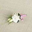 A delicately crafted wedding boutonnière lies diagonally across a softly textured, light beige fabric, the weave reminiscent of fine linen you might find in an elegant Gerrards Cross boutique. At the heart of the piece is a fully open white lisianthus bloom, its many ruffled petals forming a gentle, rounded cup that seems to glow against the neutral background. The white is crisp yet soft, with subtle shadows defining each petal, giving the flower a cloud-like lightness that feels perfect for a groom's lapel on a calm spring day. Just beside it sit two elongated, creamy buds of the same lisianthus, still closed and tinged with pale green at their tips, suggesting the quiet promise of blossoms yet to come. Threaded through the arrangement are tiny clusters of white gypsophila, or baby's breath, their minute, star-like flowers adding an airy, romantic haze that softens the composition. Slender, grass-like blades of fresh green foliage arc gracefully toward the left, introducing a sense of movement, while a broader, deeper green leaf nestles behind the main bloom, grounding the design with a hint of woodland depth. The stems at the base are neatly bound in a vivid fuchsia pink satin ribbon, tied into a small, jaunty bow that adds a playful splash of colour and a modern twist to the classic white-and-green palette. The lighting is soft and even, much like gentle natural daylight filtering through a window overlooking West Common, highlighting each detail with calm clarity and making this petite floral piece feel both intimate and celebratory-ideal for weddings, proms, or formal events across Gerrards Cross and the surrounding Chiltern villages.