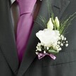 A sharply focused close-up of a groom's charcoal grey suit in Gerrards Cross, showing the left lapel dressed with an elegant white lisianthus buttonhole that perfectly echoes the rich magenta silk tie. The suit jacket appears to be fine wool with a subtle woven texture, its lapel crisp and structured, framing the floral detail with clean lines. Nestled against the fabric, a fully opened white lisianthus bloom shows off its soft, ruffled petals, layered like fine tissue, creating a gentle focal point of purity and calm. Just above, two slender ivory buds lean in, still closed and smooth, suggesting blossoms that will unfurl later, like the quiet anticipation before the vows at a wedding ceremony in one of the churches near East Common. Surrounding the main flower is a light cloud of baby's breath, each tiny white floret airy and delicate, giving the buttonhole a romantic, almost mist-like halo. Slim blades of fresh greenery arc outward, adding movement and a natural, garden-picked feel that contrasts beautifully with the tailored charcoal fabric. At the base, a narrow mauve ribbon is wrapped and tied in a neat bow, its sheen catching the light and gently echoing the deeper tones of the tie. The overall look feels refined yet heartfelt, as if prepared by an experienced local florist who understands how a small detail on a lapel can carry the quiet emotions of a wedding day, from intimate civil ceremonies near the railway station to grand celebrations in country venues just outside Gerrards Cross.