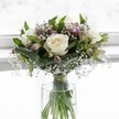 In this softly lit scene, a compact, elegant bouquet of creamy white roses and delicate accent flowers rests in a simple cylindrical glass vase, positioned against a luminous, blurred white background that feels like a bright Gerrards Cross morning. Three pristine white roses, fully open with velvety petals spiralling towards subtle ivory centres, form the calm heart of the design. Around them, rich emerald foliage curves and folds, its broad leaves gently cupping the blooms and giving the arrangement a sense of structure and grace, much like the manicured greenery you might see around West Common. Threaded between the roses are fine, cloud-like sprays of gypsophila, or baby's breath, their tiny white blossoms hovering on slender stems and softening every edge with an almost misty lightness. Nestled a little deeper in the design, partially open alstroemeria buds introduce whisper-soft touches of mauve and blush pink, their future colour promised rather than fully revealed, suggesting quiet romance and thoughtful intention. The clear vase reveals neatly gathered stems, cleanly trimmed and tied with a discreet ribbon just above the waterline, a sign of careful, professional floristry. Natural daylight sweeps across the flowers, catching highlights on each rose petal and glancing off the water's surface, creating a tranquil, almost devotional mood. The bouquet feels perfectly suited to a peaceful wedding, a christening near the local church, or a gentle condolence gesture delivered to a home in Gerrards Cross, offering comfort, purity, and understated sophistication with every stem thoughtfully placed.