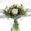 A soft, contemplative bouquet is arranged in a clear, straight-sided glass vase, its delicate colours and textures evoking the quiet stillness of a gentle farewell. At the centre, white roses open fully, each bloom a careful study in texture, with velvety petals unfolding in creamy layers that seem to glow even in the soft light. Threads of gypsophila weave around them, thousands of tiny white blossoms creating a mist-like halo that blurs the boundaries between each flower, giving the arrangement an almost weightless feel. Rising through this pale cloud are alstroemeria stems, their buds pointed and slender, some just beginning to part and reveal petals tipped with pale lavender and muted purple, touched with fine stripes that add subtle character rather than bold colour. Framing the flowers is a collar of deep green, broad, tropical-style leaves, glossy and smooth, gently wrapped to hold the bouquet in a rounded, hand-tied form that speaks of professional craftsmanship. Additional narrow leaves and soft green stems pierce the design, lending height and a sense of living movement without disturbing its calm symmetry. The vase itself is filled with clear water, through which you can see the clean-cut stems, neatly bundled and secured just above the waterline, a testament to their freshness. Diffused light, reminiscent of a quiet afternoon filtering through a window near Bulstrode Way or along Packhorse Road in Gerrards Cross, bathes the flowers in a tranquil glow. The mood is one of solace and remembrance, a comforting floral expression well suited to sympathy deliveries, designed to bring a measure of peace to those who receive it.