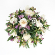 Seen from directly overhead, this hand-crafted sympathy bouquet forms a soft, rounded shape set against a pristine white background, as if it is resting on a bright table in a peaceful Gerrards Cross sitting room. The centre is composed of several full, creamy white roses, their petals unfolding in perfect spirals that progress from cool ivory edges to warm, almost peachy cores. Some blooms are wide open, allowing you to sense their velvety texture and gentle fragrance, while a few remain more tightly cupped, symbolising emotions still held close. Threaded between the roses are slender purple alstroemeria buds, most of them still in their early, closed stages. Their outer petals are painted in gradients of rich lavender and deep purple at the tips, softening into fresh green and hints of pink as they approach the stems, suggesting colour yet to fully emerge. Light clusters of delicate white baby's breath float among the larger flowers, each tiny blossom like a fleck of cloud or a whispered breath, softening the arrangement and lending it an airy, almost weightless feel. Around the perimeter, broad, deep green leaves with a natural sheen cradle the blooms, creating a lush background that enhances the whites and purples and defines the bouquet's gentle, spherical silhouette. The overall look is calm, dignified, and soothing, calling to mind quiet moments of reflection, perhaps after a walk near the local churches or green spaces in Gerrards Cross. It feels like an arrangement chosen with care to convey sympathy, purity, and a comforting presence when silence speaks louder than words.