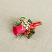 A richly detailed boutonniere is laid against a softly textured, light beige linen background, the sort of fabric you might see on a rustic table in a Gerrards Cross country-style reception venue. At its heart sits a single deep fuchsia pink rose, the petals tightly coiled in the centre and gradually unfurling into a velvety spiral, suggesting both freshness and quiet anticipation. Around the base of this rose, tiny white blooms of baby's breath cluster like a gentle cloud, their minute petals creating a delicate, airy halo that softens the intensity of the pink. Threaded between these white sprigs are glossy red hypericum berries, perfectly round and polished, lending a festive pop of crimson that hints at winter weddings or autumn celebrations around West Common and the leafy lanes nearby. Wisps of fine, fern-like greenery extend from the composition, adding feathery movement, while broader dark green leaves are carefully curled and tucked in to give structure, depth, and a sense of subtle drama. The stems are bound neatly in a rich red satin ribbon, wrapped smoothly and tied into a small, elegant bow that glints slightly in the gentle, natural light, evoking the feeling of an intimate ceremony at a local Gerrards Cross church or registry office. The boutonniere is photographed from a slightly elevated angle, which allows every curve of petal, sheen of berry, and texture of foliage to be appreciated, as if a florist were holding it up for a client to inspect before pinning it to a lapel. It feels ideal for a groom or groomsman who wants a classic, romantic accent with a modern, vibrant twist, the sort of handcrafted detail a local florist would create with care for weddings and formal events across Gerrards Cross and the surrounding Buckinghamshire villages.