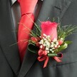 A close-up, high-angle photograph reveals an exquisitely crafted buttonhole pinned neatly to the left lapel of a charcoal grey suit jacket, the kind you might see on a groom stepping out of a classic Gerrards Cross church on a bright wedding morning. At the heart of the boutonnière sits a single, perfectly formed deep red rose bud, its velvety petals tightly furled yet full of promise, standing upright with quiet confidence. Around the base of the rose, tiny white blooms reminiscent of baby's breath create a soft, cloud-like halo, their airy texture adding lightness against the dark fabric of the suit. Nestled among these petite flowers are small, glossy red berries, their rounded shapes echoing the rose's rich crimson tone and adding a subtle, almost jewel-like sparkle in the gentle indoor light. Slender, grass-like blades of greenery arch outwards from behind, while broader, darker green leaves are wrapped snugly at the base, giving structure and balance. A narrow red satin ribbon, carefully tied into a neat bow, secures the stems and links the red accents together with a sense of polished refinement. Just beneath the lapel, a bright red tie in smooth satin or silk is visible, its perfectly formed knot aligned with the buttonhole and reinforcing the cohesive, romantic colour palette. A lightly patterned dress shirt in pale grey or white peeks through, its fine checks or houndstooth weave adding a discreet layer of texture. This detailed image captures the care and craftsmanship a local florist in Gerrards Cross brings to men's wedding flowers, ideal for illustrating formal attire, boutonnière styling, or special event dressing for celebrations across the town and nearby Chiltern villages.