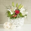 A beautifully composed floral display is shown as if set for a wedding reception or special dinner party in Gerrards Cross, perhaps on a long table near West Common. At its heart is a distinctive, round white vase with an intricate open-weave design, resembling white wicker or delicately tangled twigs, which adds a sculptural, contemporary touch. The vase is filled to overflowing with a generous bouquet, dominated by elegant white lilies. Several lilies are already in full bloom, their broad petals curving outward in smooth, creamy arcs around golden stamens, while other buds remain tightly closed, their green casings pointing upwards and signalling freshness. Nestled close to these lilies are velvety red roses, one central bloom especially striking, its deep crimson petals spiralling inwards to a perfect, romantic centre. Bright green, ball-shaped flowers-likely green dianthus or chrysanthemum pom-poms-sit between the lilies and roses, introducing a lively texture and a vivid dash of green that enlivens the whole composition. Fine clusters of white baby's breath are threaded gently among the larger blooms, giving a light, airy feel, while sprigs of small red berries bring a touch of seasonal festivity suitable for winter weddings or Christmas gatherings. A rich mix of foliage, from slender, feathery stems to broader, darker leaves, frames the flowers, creating depth and a natural, garden-inspired look. To the left of the main vase, two decorative spheres made from pale, woven strands echo the vase's texture and rest quietly on the white surface. Behind, a softly textured neutral wall in beige or light grey creates a calm backdrop, drawing attention to the crisp whites, deep reds, and fresh greens that would enchant guests at any table in Gerrards Cross.
