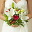 In this photograph, a hand-tied bridal bouquet is cradled against a lace wedding dress, its details captured with the same care a florist might show when preparing flowers for a ceremony in one of Gerrards Cross's elegant churches. The focal point is a trio of open white lilies, their star-shaped petals creamy and luminous, each bloom revealing a delicate centre of pollen-tipped stamens that hints at a gentle, powdery scent. Nestled beside and between them are velvety red roses, petals curling tightly toward the heart, their crimson tone rich and passionate against the purity of the lilies. Tiny sprigs of gypsophila, or baby's breath, scatter throughout the bouquet like soft, white confetti, lightening the composition and adding an airy texture. Bright red berries, most likely hypericum, cluster in small bursts that feel cheerful and contemporary, while tufts of moss-like green dianthus and rounded moss balls introduce a playful, woodland character reminiscent of shaded paths around Austenwood. Various shades of foliage-from glossy, deep green leaves to softer, muted greens-form a lush backdrop, creating dimension and framing the blooms with a sense of abundance. Fine, arching grasses extend beyond the main body of the bouquet, giving it an almost ethereal outline, as if the flowers are gently floating. The overall effect is one of refined romance and considered craftsmanship: classic wedding flowers arranged with modern sensitivity, perfectly suited to a bride walking through Gerrards Cross, whether she is emerging from the registry office or stepping into a reception at a nearby country-style venue.