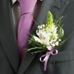 A close-up, high-angle photograph captures the upper torso of a man dressed for a formal event in Gerrards Cross, his attire centred around a charcoal grey suit jacket with sharply tailored lapels and a softly structured shoulder line. Beneath the open jacket, a crisp, pale dress shirt with a faint grid pattern peeks through, its cool tones perfectly offset by a smooth lavender necktie that runs in a clean vertical line down the chest. Pinned to the left lapel, a meticulously crafted wedding buttonhole becomes the focal point: a light green spire of unopened buds, suggesting hyacinth or Muscari, rises elegantly, adding height and a sense of fresh anticipation. Around this central stem, white Alstroemeria blooms open gently, their petals marked with fine maroon and purple streaks and golden stamens that glimmer softly in the ambient light. Tiny clusters of white baby's breath, or Gypsophila, nestle between the larger flowers, creating an airy, cloud-like halo. Slender, grass-like blades and broader, glossy green leaves frame the composition, offering movement and a vivid contrast to the dark jacket and pale shirt. At the base, a lilac satin ribbon is tied into a neat, compact bow, perfectly echoing the lavender tie and drawing the elements together with quiet harmony. The overall mood is one of poised celebration, evoking a groom or groomsman getting ready in a calm room before a ceremony at a local church or venue near East Common or West Common, the fresh fragrance of stems and petals grounding the emotion of the moment in something tender and real.
