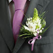 This image focuses closely on a man's formal attire, where the artistry of the floral work is just as important as the cut of the suit. A dark charcoal jacket fills the frame, its lapel clean and structured, hinting at a thoughtfully chosen outfit for a wedding at a Gerrards Cross church or an evening event near the station. At the centre of the composition, pinned carefully to the left lapel, sits a striking boutonniere made up of crisp white alstroemeria blossoms, their petals gently flared. Fine reddish-brown and violet freckles decorate the inner petals, leading the eye towards warm yellow centres where stamens add delicate detail. A tapering green spike, reminiscent of an unopened tuberose stem, stands proudly from the heart of the design, bringing a fresh, vertical energy that suggests new beginnings. Around it, clusters of white baby's breath form a soft haze, while fine, grassy fronds and rich, deep-green leaves create layers of texture, from feathery to smooth. Just beside the flowers, a lavender tie lies perfectly straight against a subtle, patterned white shirt; its silky surface reflects the light, and its colour is mirrored in the lilac satin ribbon wound at the base of the boutonniere and finished in a precise bow. The lighting is gentle, almost like natural daylight streaming into a Gerrards Cross home before a ceremony, highlighting the contrast between cool greys, luminous whites, and fresh greens. Everything about the arrangement feels balanced and intentional, from the scale of the blooms to the placement on the lapel, conveying a quiet confidence and a sense of anticipation for whatever milestone moment the wearer is about to step into.