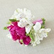 A compact, hand-crafted floral arrangement lies horizontally on a softly textured, light beige fabric that resembles natural linen, the neutral backdrop gently cradling every detail of the flowers. At the heart of the design is a lush, fully opened peony in rich fuchsia-magenta tones, its many ruffled petals layered so densely that they form a velvety, cushion-like bloom, almost overflowing with colour and softness. Around this central peony, several white alstroemeria blossoms unfold with quiet elegance, their six slender petals tipped in creamy white and delicately marked with fine maroon and deep red streaks that look like careful brushstrokes. These Peruvian lilies curve slightly outward, their speckled throats adding a sense of movement and intricacy. Threaded between the main flowers are slim, spiky green buds and florets, possibly young gladiolus or similar filler stems, offering fresh, bright green accents and a linear contrast to the roundness of the peony. The stems are neatly aligned and wrapped in a bright pink satin ribbon, its glossy surface catching the light and perfectly echoing the bold hue of the central bloom. The lighting feels gentle and natural, as if the arrangement has been placed near a window in a calm Gerrards Cross home, perhaps just prepared for a wedding at the local church or a celebration in a garden near East Common. The overall mood is joyful yet refined, a small but striking piece that could serve as a boutonnière, bridesmaid posy, or corsage, capturing the romance of a spring or summer day in Gerrards Cross with its harmonious blend of fuchsia, white, and fresh green.